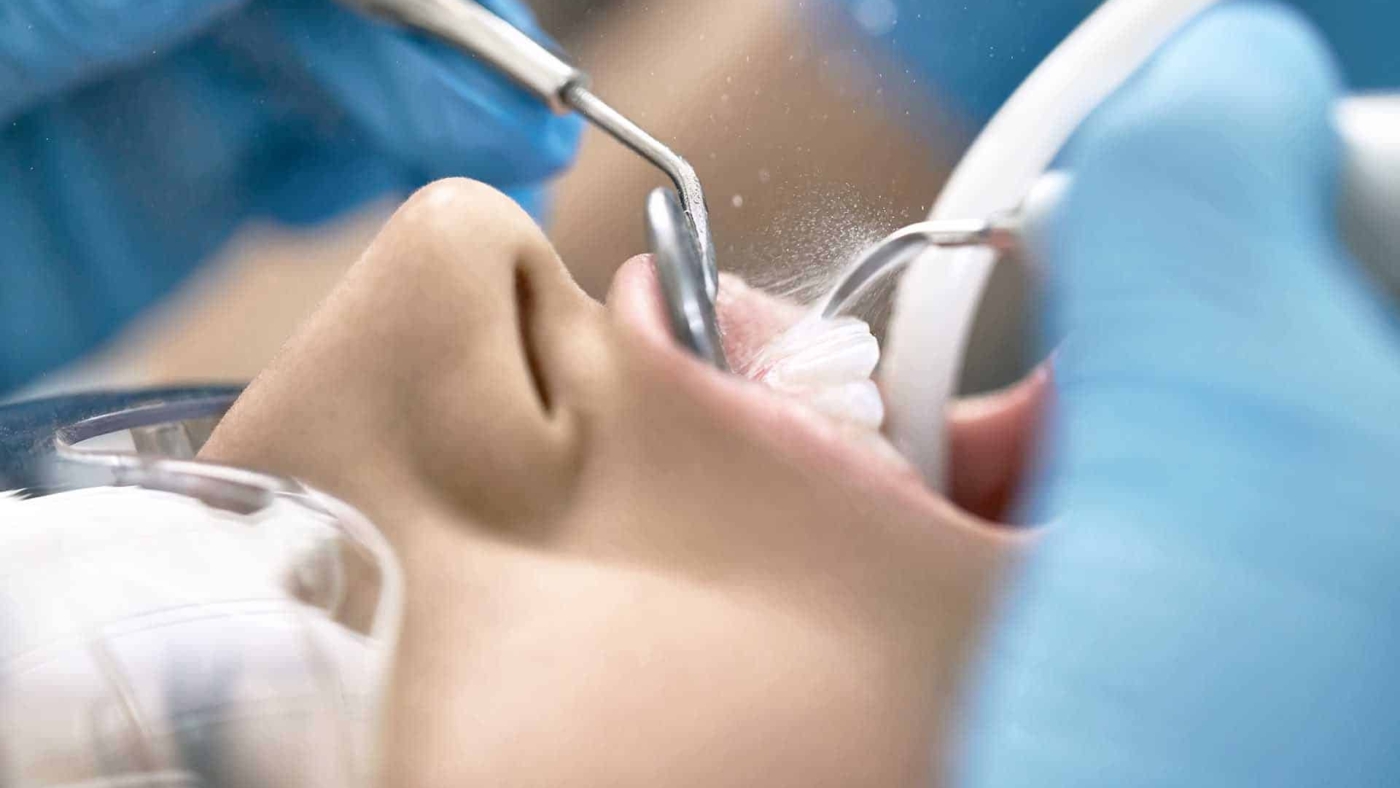 Female patient in protective glasses in a dental clinic. Dentist in blue latex gloves is cleaning her teeth with a help of a water jet, dental mirror and a saliva ejector. Closeup horizontal photo.